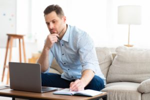 Pensive man using laptop at home and writing notes